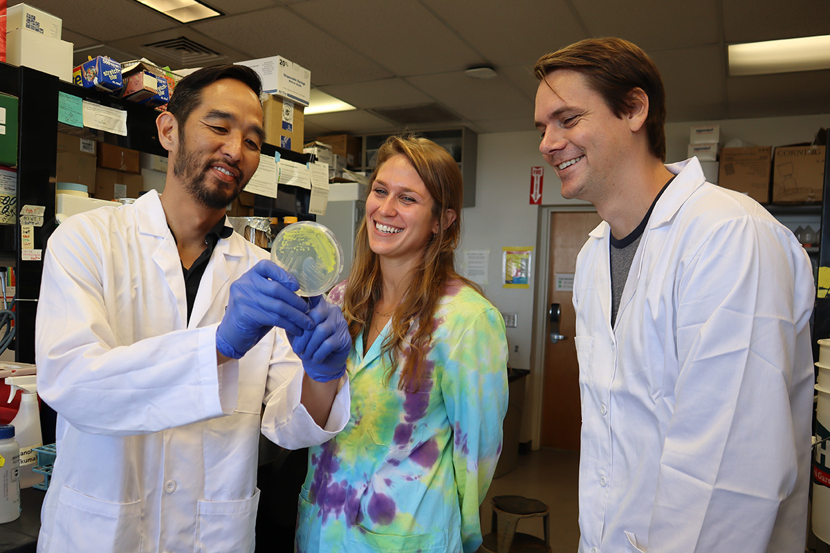 Metamorphotech trio of scientists in lab coats looking at Petri dish, smiling